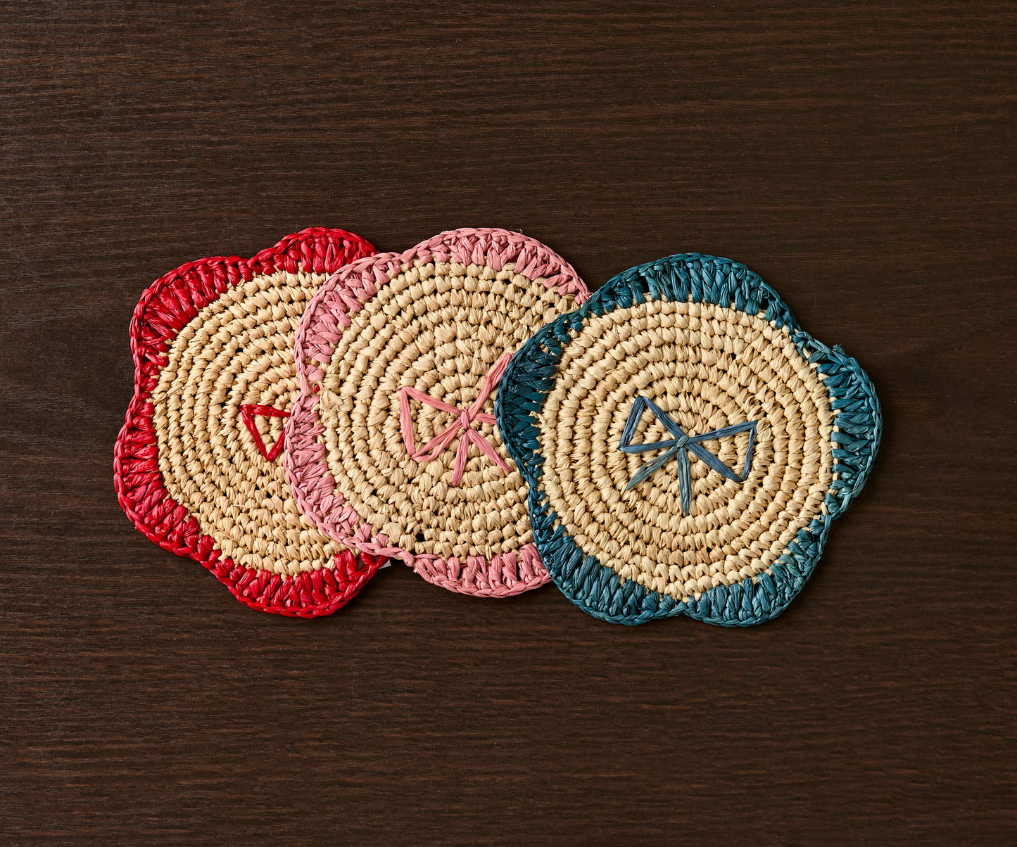 Handwoven raffia fringe coasters in red, pink, and teal, featuring a floral design on a dark wooden table.