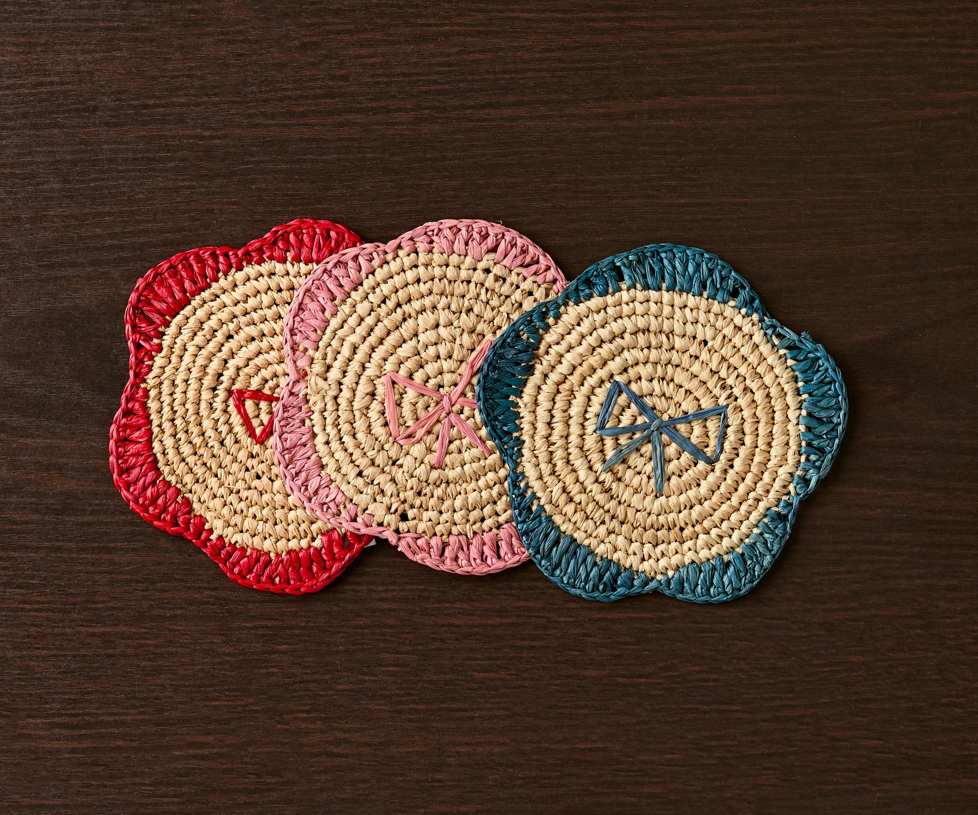Handwoven raffia fringe coasters in red, pink, and teal, featuring a floral design on a dark wooden table.