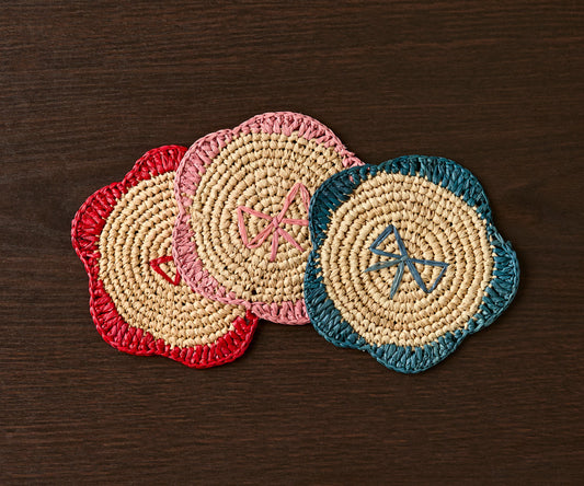 Handwoven raffia fringe coasters in red, pink, and teal, featuring a floral design on a dark wooden surface.
