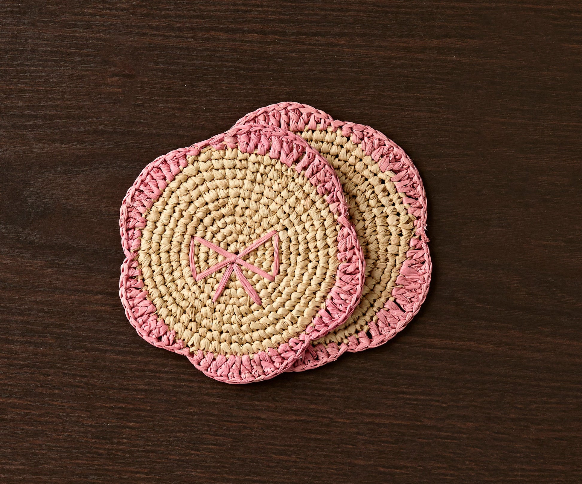 Handwoven raffia fringe coaster with a pink edge and bow design, placed on a dark wooden surface.