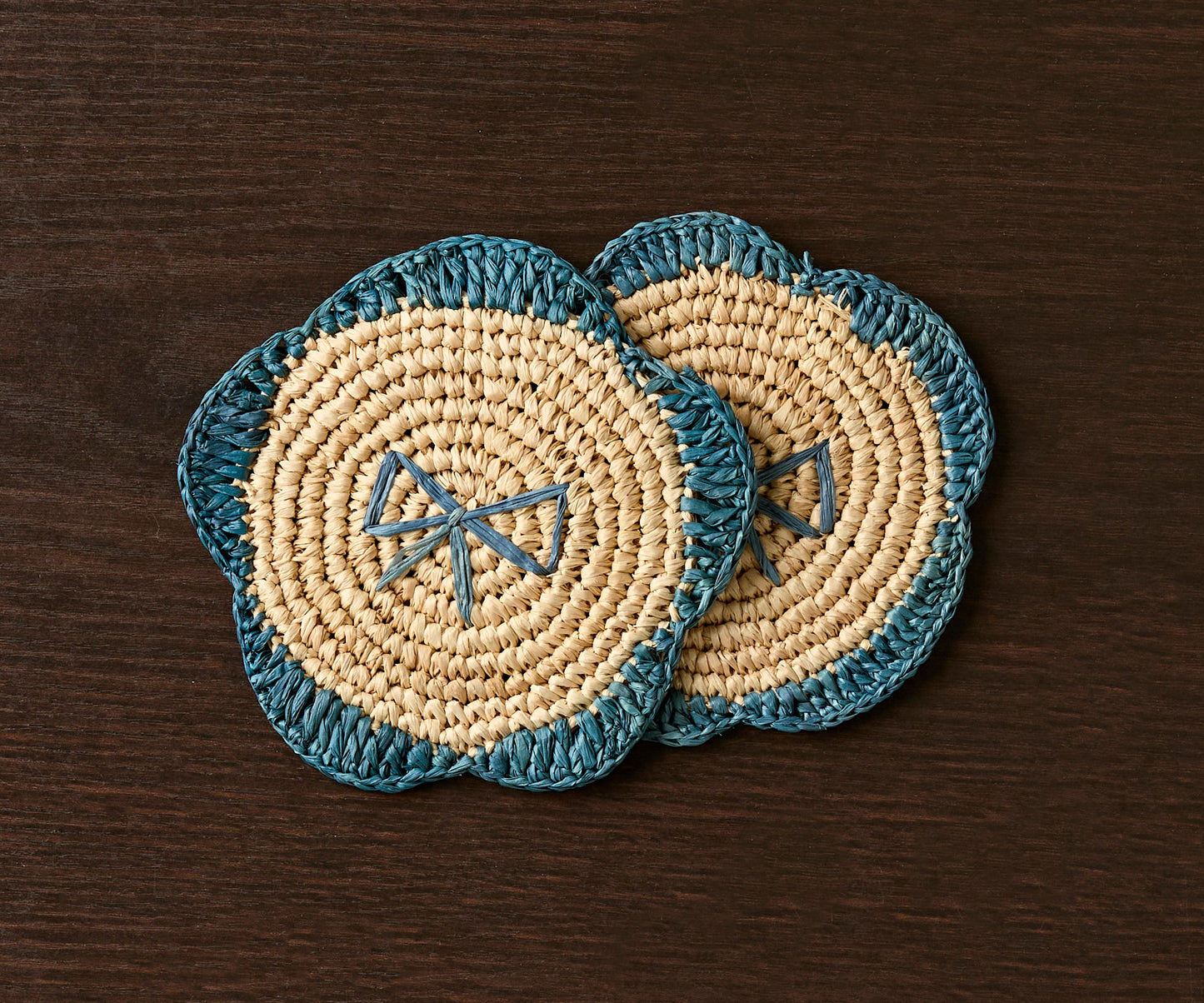 Handwoven raffia fringe coasters in natural and teal colors, featuring a floral design on a dark wooden surface.