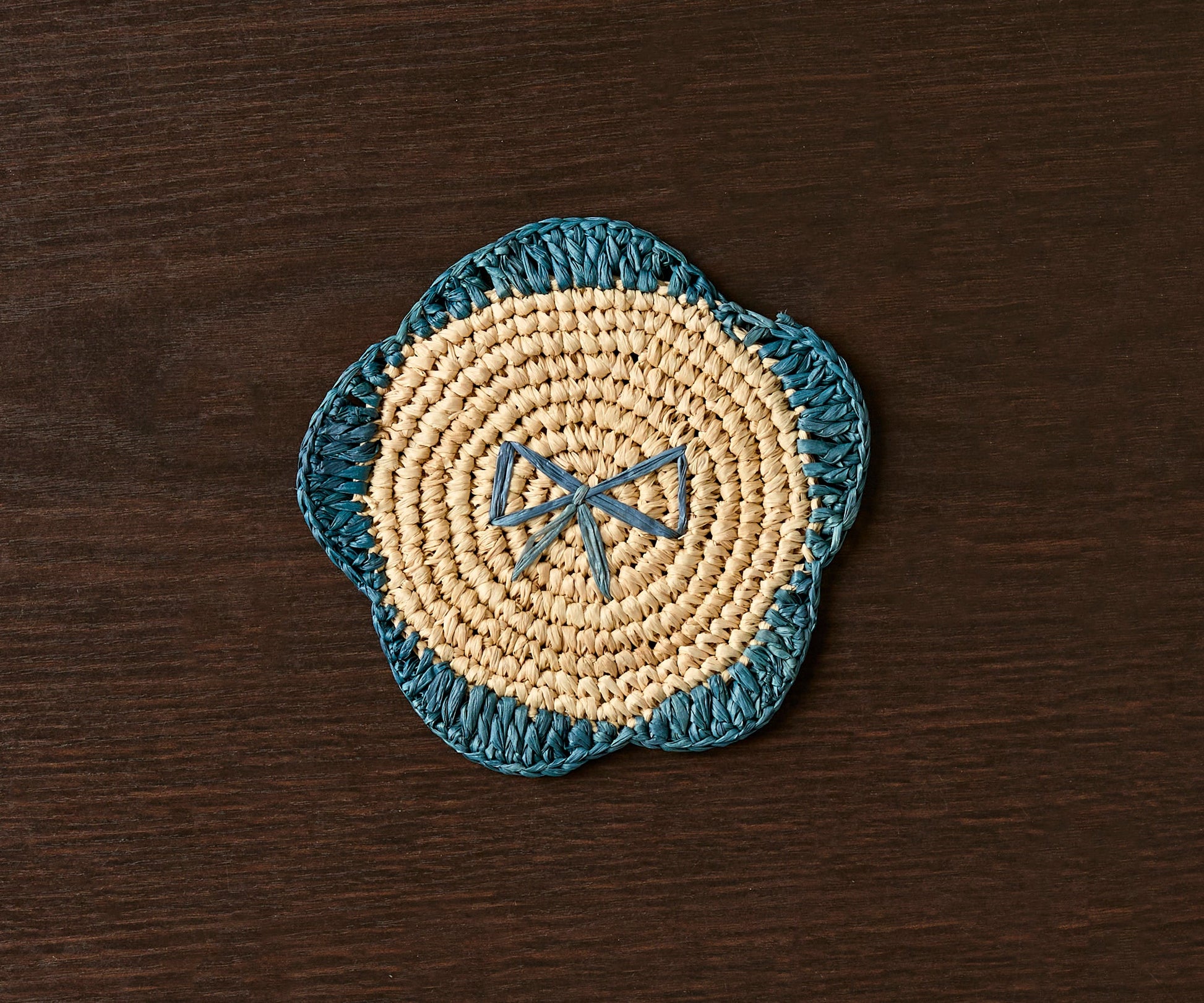 Handwoven raffia fringe coaster in a flower shape, featuring natural raffia and teal accents on a dark wooden surface.