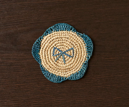 Handwoven raffia fringe coaster in a flower shape, featuring natural raffia and teal accents on a dark wooden surface.