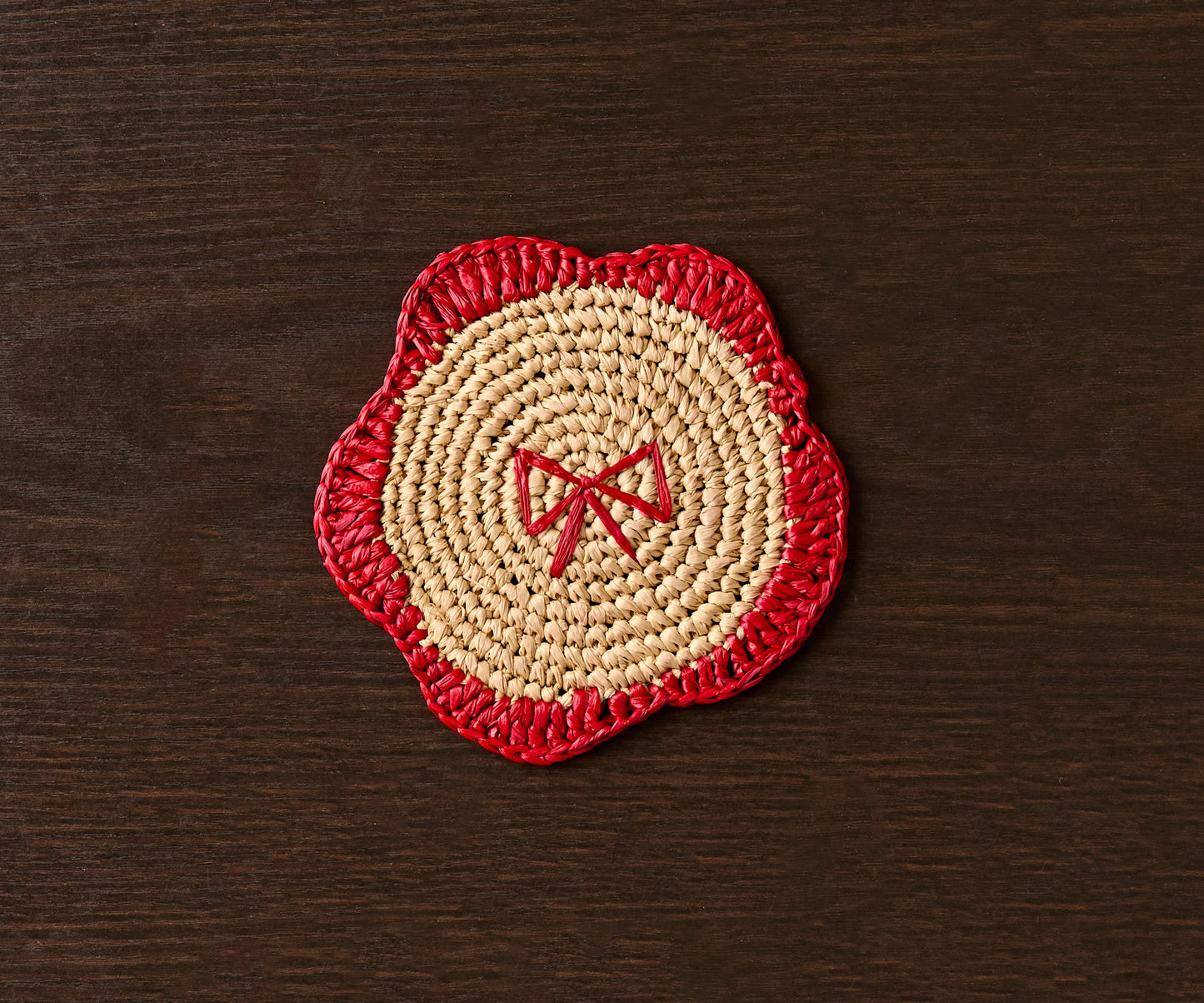 Handwoven raffia coaster with a red scalloped edge and central bow design on a dark wooden table.