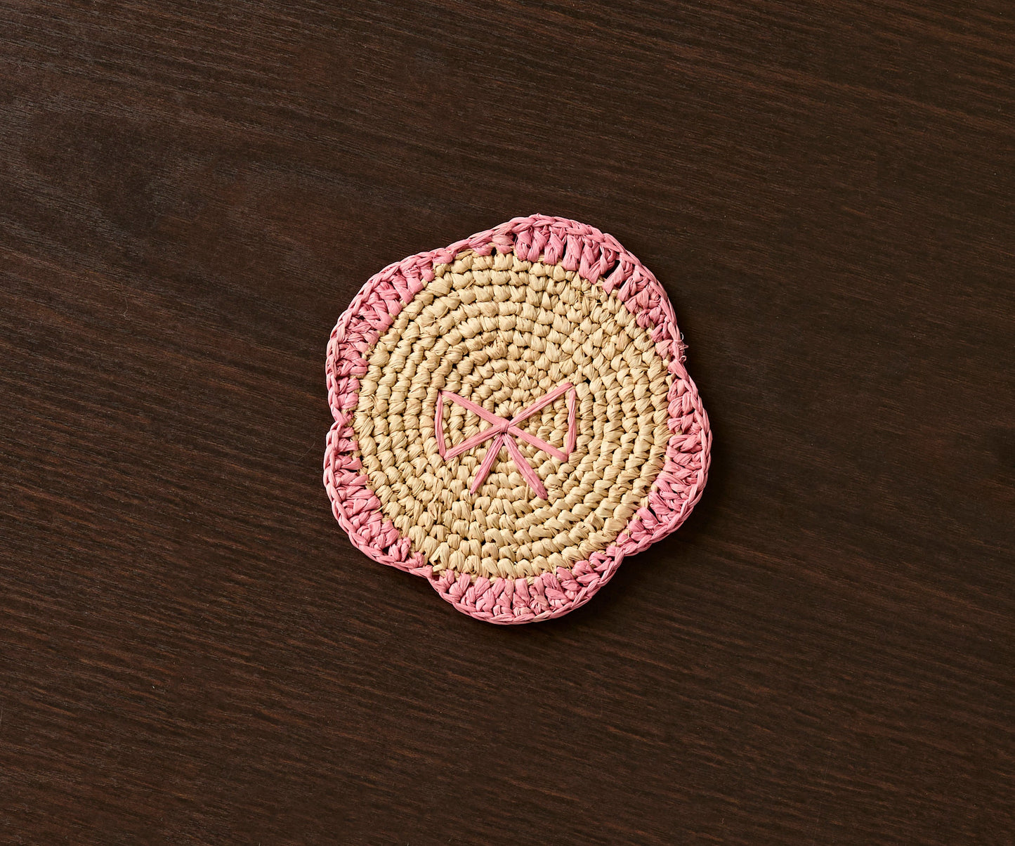 Handwoven raffia coaster with a pink fringe and bow design on a dark wooden surface.