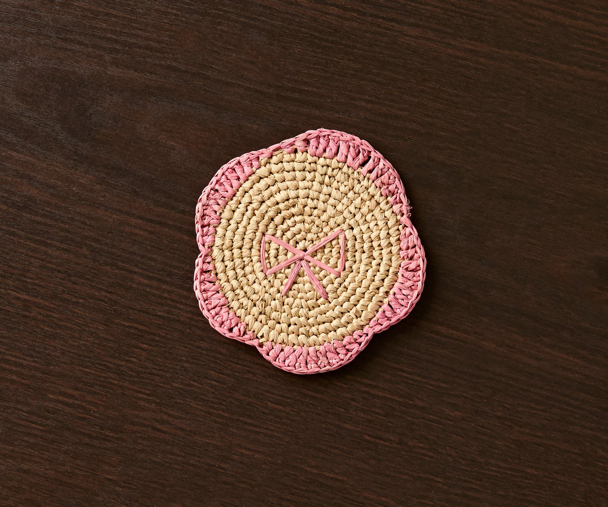 Handwoven raffia coaster with a pink fringe and bow design on a dark wooden surface.