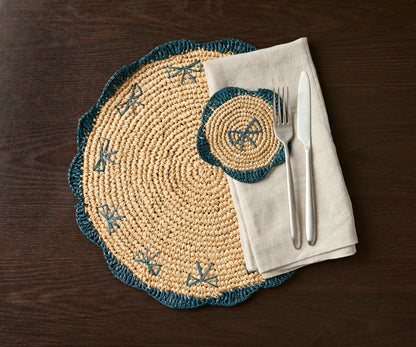 Handwoven raffia fringe coaster in natural tones with teal accents, placed on a dark wooden table with utensils.