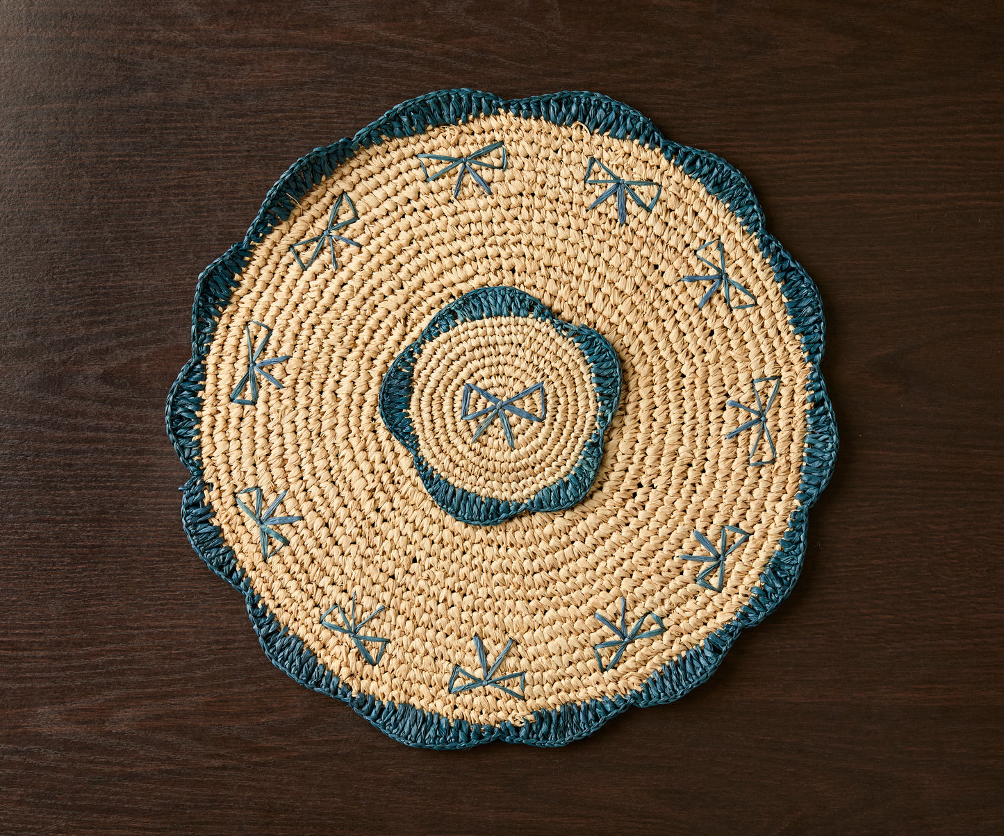 Handwoven raffia fringe coaster with blue accents, featuring a circular design and intricate patterns, on a dark wooden su...