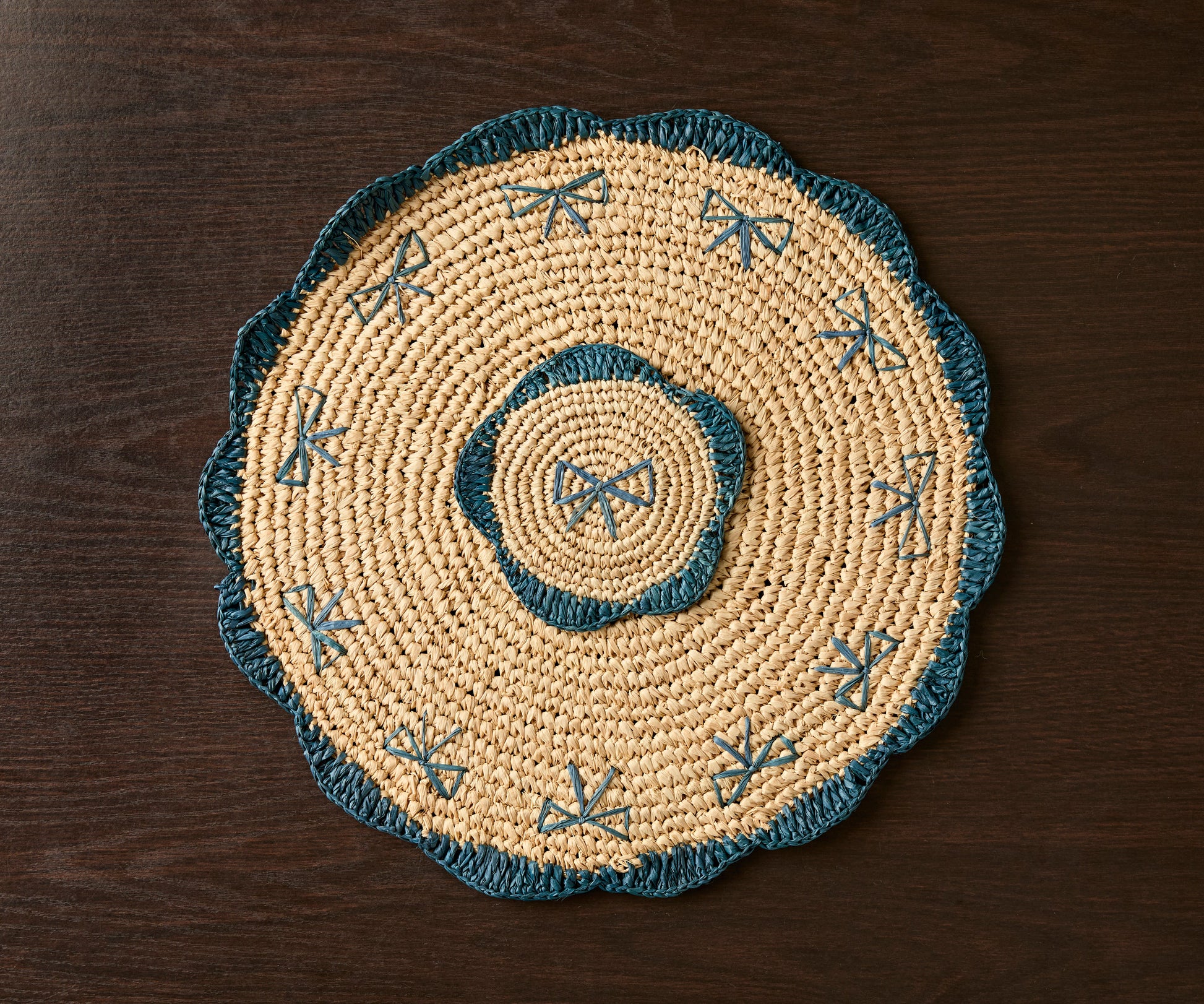 Handwoven raffia fringe coaster with blue accents, featuring a circular design and intricate patterns, on a dark wooden su...