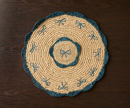 Handwoven raffia fringe coaster with blue accents, featuring a circular design and intricate patterns, on a dark wooden su...