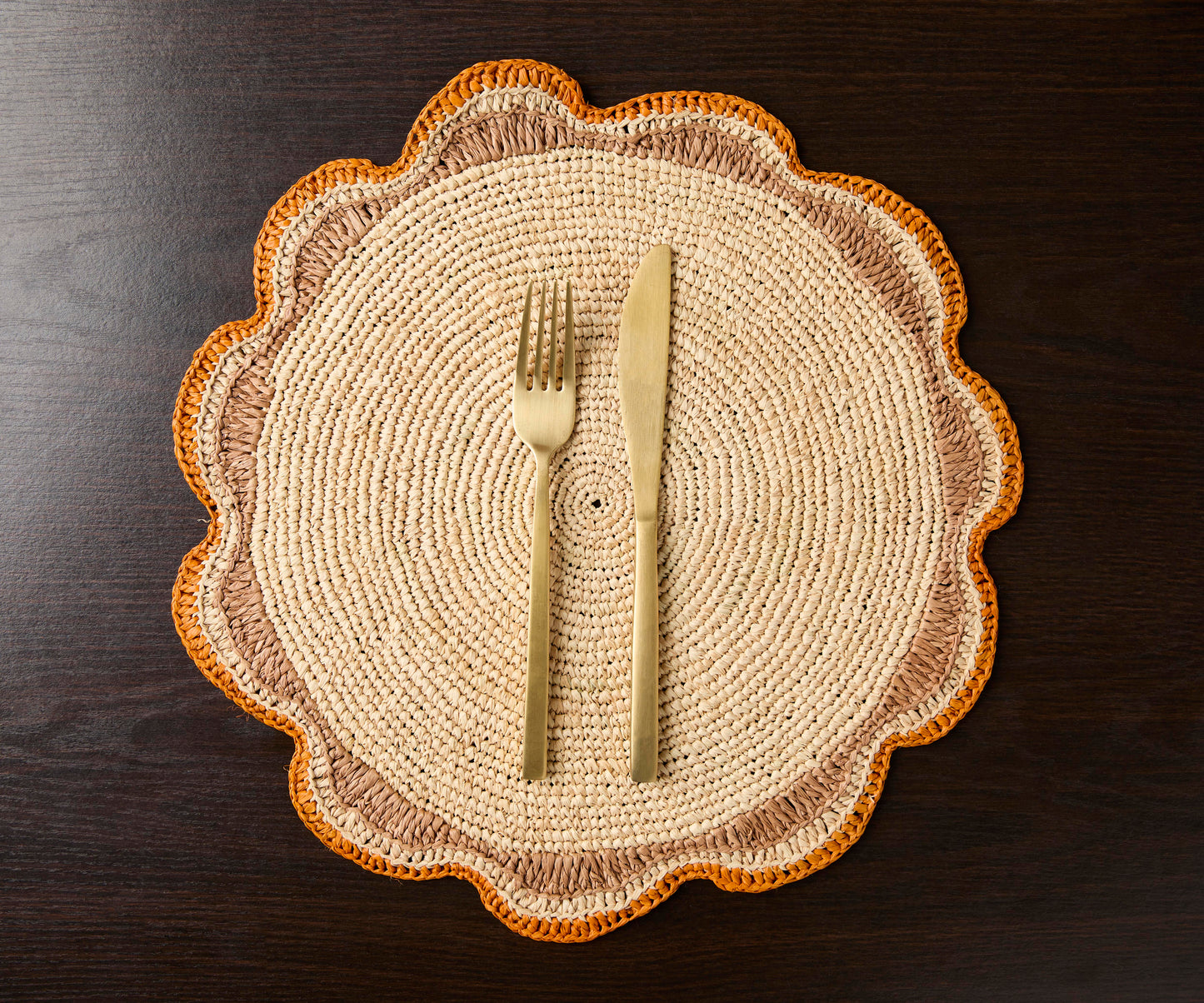 Round raffia woven placemat with scalloped edges, featuring beige and orange tones, accompanied by a fork and knife.