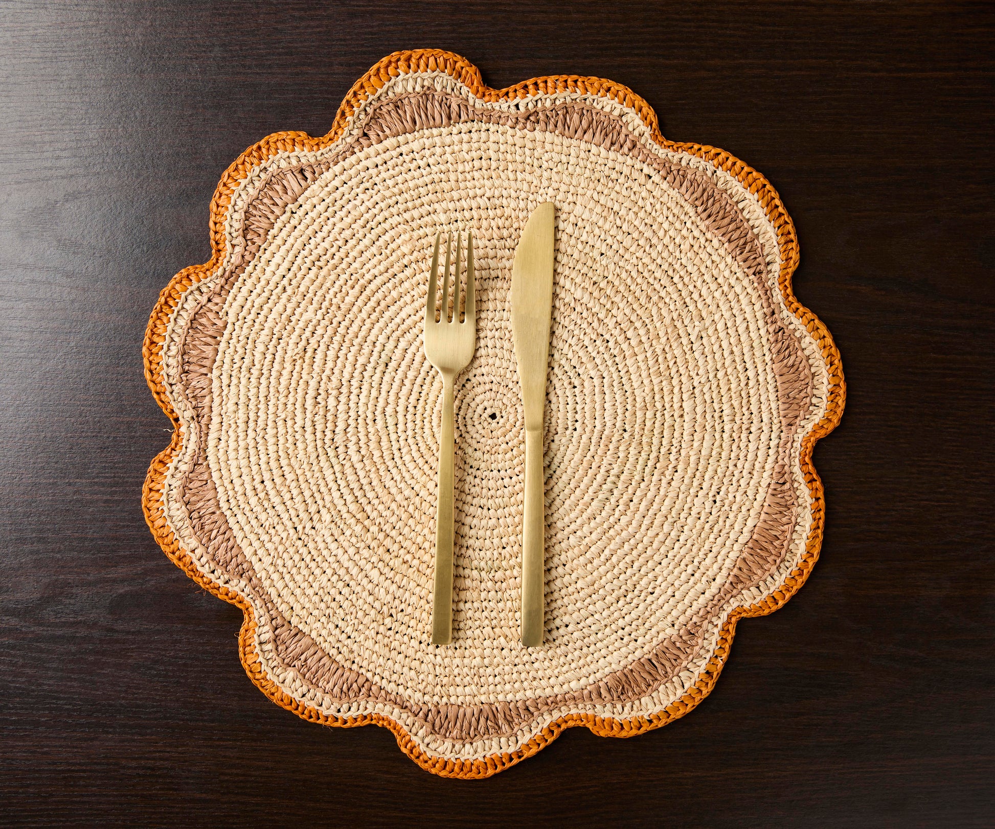 Round raffia woven placemat with scalloped edges, featuring beige and orange tones, accompanied by a fork and knife.