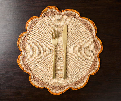 Round raffia woven placemat with scalloped edges, featuring beige and orange tones, accompanied by a fork and knife.