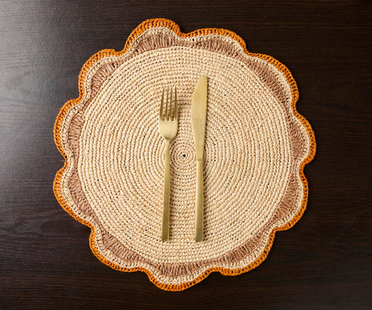 Round raffia woven placemat with scalloped edges, featuring beige and orange tones, accompanied by a fork and knife.