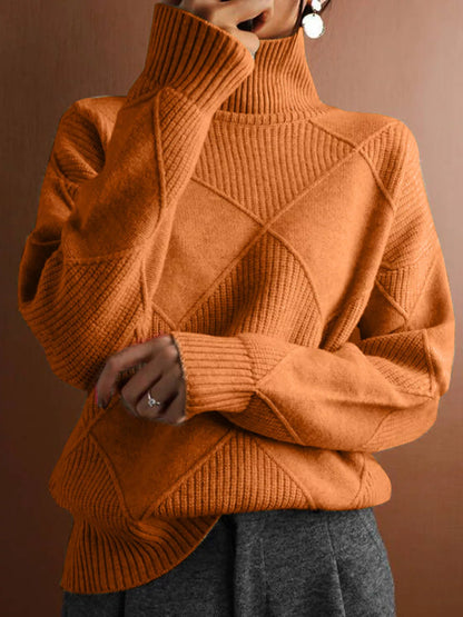 A person wearing a warm rust-orange ribbed turtleneck sweater with diamond-shaped knit paneling on the front, paired with gray pants, against a solid brown background.