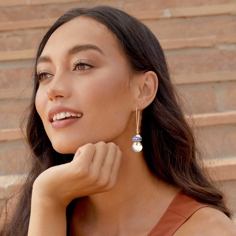 A woman with long dark hair wearing a dangling earring with a gold-toned top, a small blue stone, and a white pearl drop, posed against a brick wall.