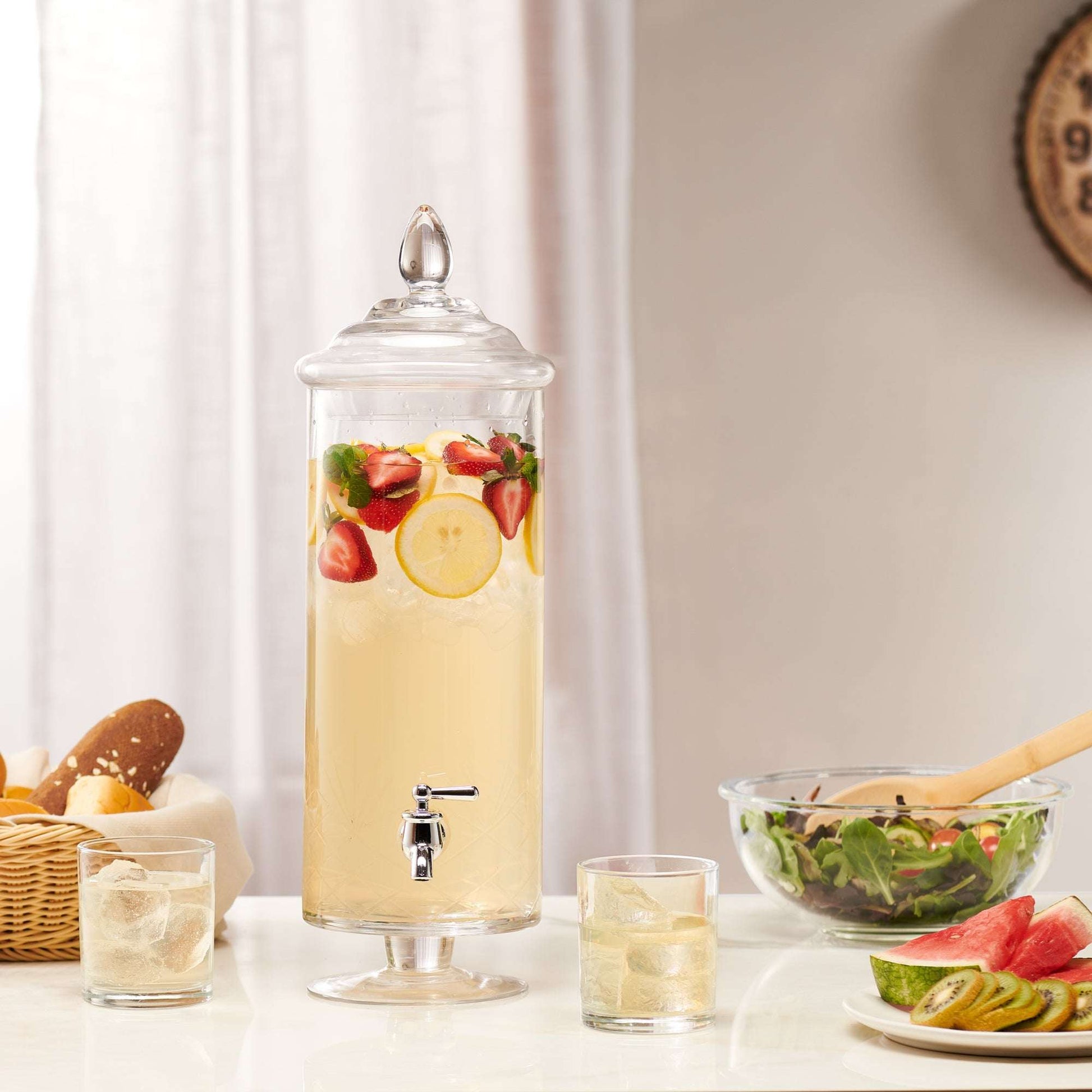 Tall glass beverage dispenser with a metal spigot, filled with lemon and strawberry water on a table with two glasses.
