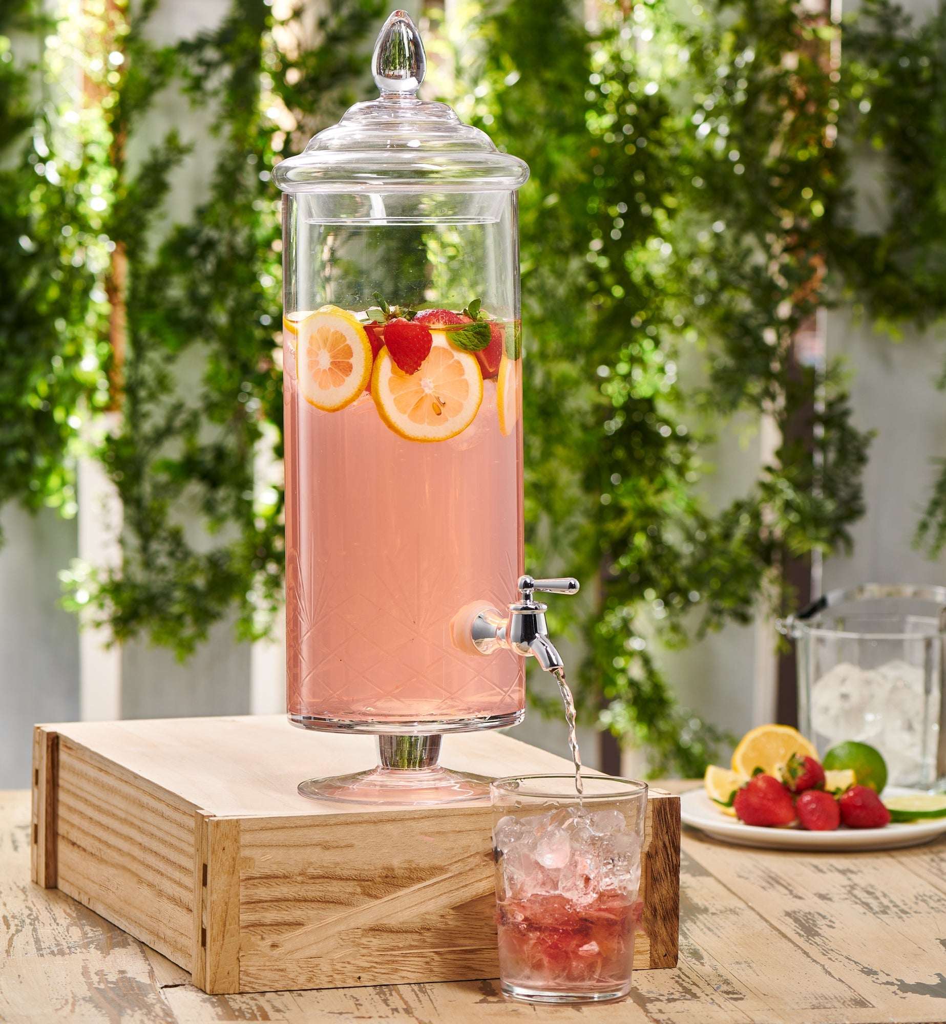 Tall glass beverage dispenser with a metal spigot on a wood crate, filled with pink liquid, lemon slices, and strawberries.