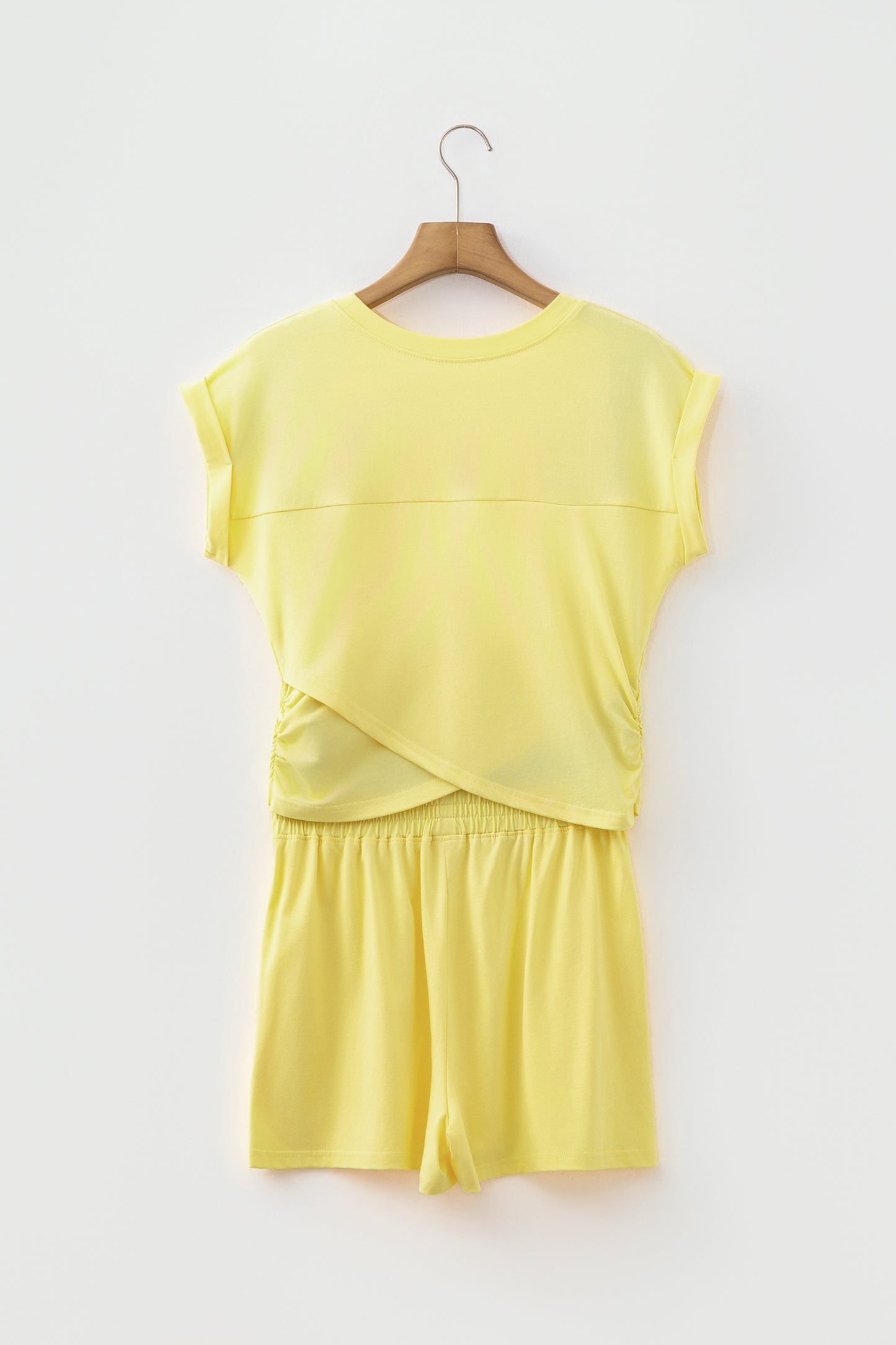 A two-piece matching lounge set in pale yellow, featuring a short-sleeve top with a wrap-style front and matching shorts, hanging on a wooden hanger against a plain white wall.