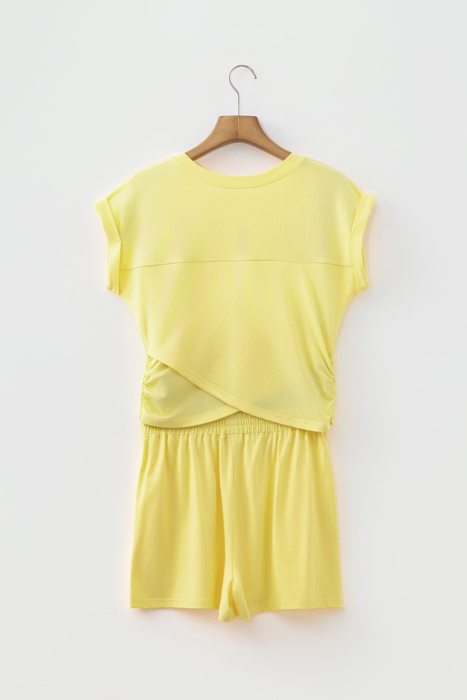 A two-piece matching lounge set in pale yellow, featuring a short-sleeve top with a wrap-style front and matching shorts, hanging on a wooden hanger against a plain white wall.
