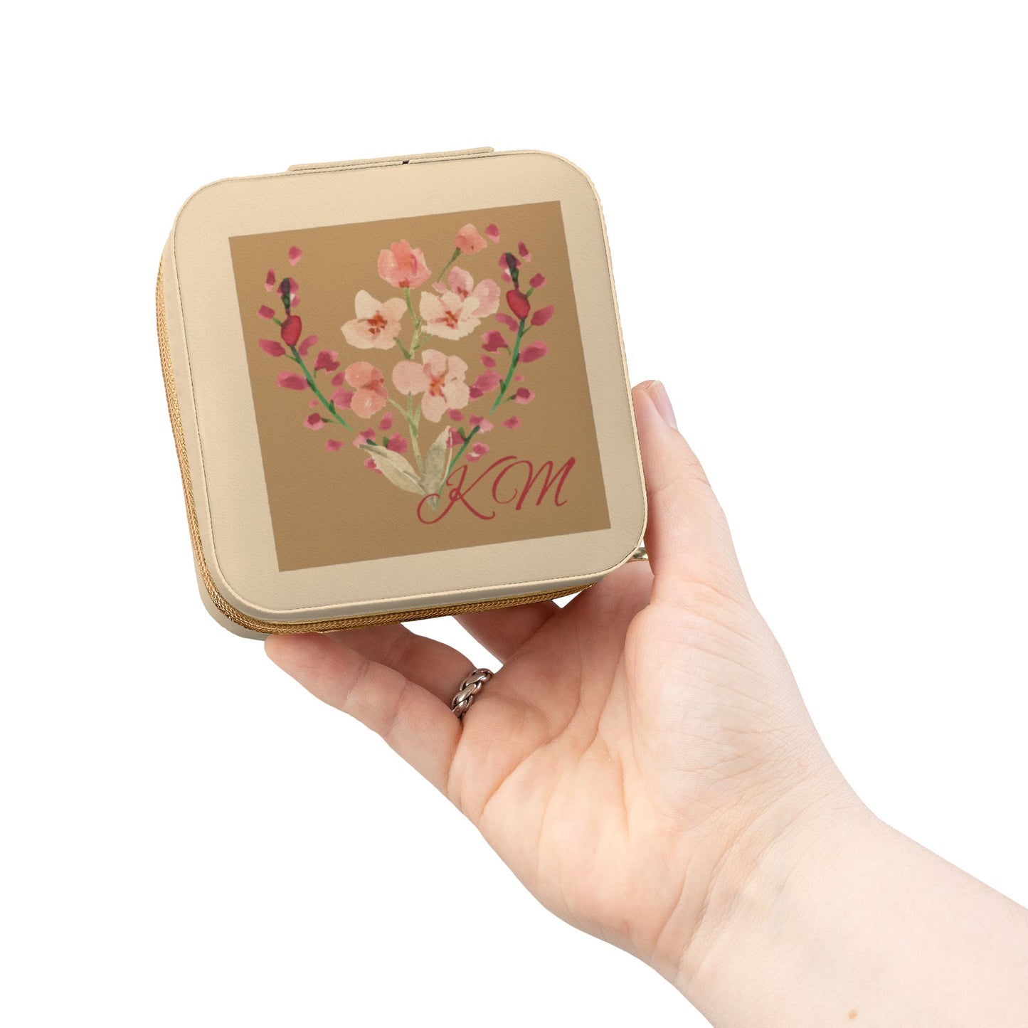 A hand holding a small square compact case with rounded corners. The case is beige with a central floral illustration in pink, peach, and green, and the initials 'KM' in red script.