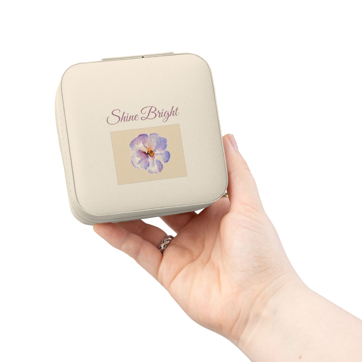 A hand holding a small square cream-colored cosmetic case with rounded corners. The case features the words 'Shine Bright' in a cursive font above a small illustration of a purple flower.
