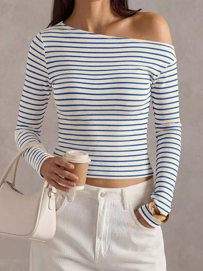 A woman wearing a blue and white horizontally striped off-the-shoulder long-sleeve crop top, paired with white pants and holding a beige handbag and a paper coffee cup against a neutral gray background.