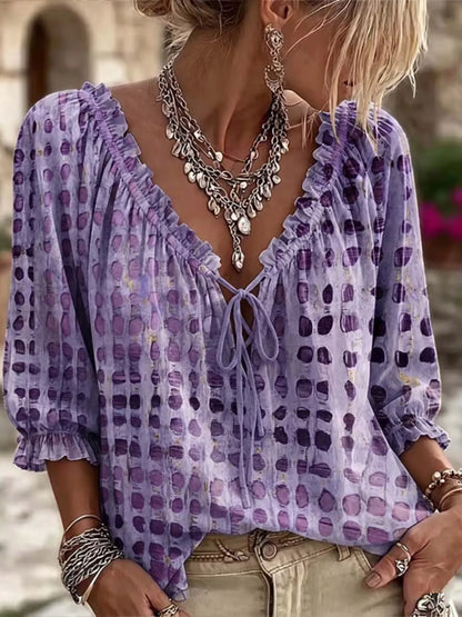 A woman wearing a lavender purple tie-front blouse with a watercolor-style polka dot pattern, paired with beige pants and layered silver jewelry.