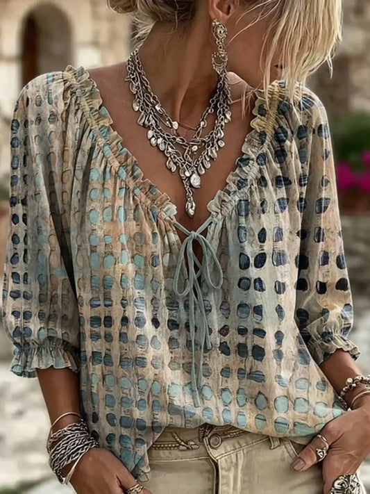 A woman wearing a bohemian-style long-sleeve blouse with a tie-neck and an abstract spotted pattern in beige, light blue, and dark navy, styled with layered silver necklaces and bracelets.