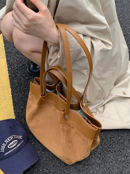 A brown leather tote bag with double shoulder straps, held by a person wearing a beige shirt. A navy blue baseball cap is visible in the bottom-left corner on dark asphalt.