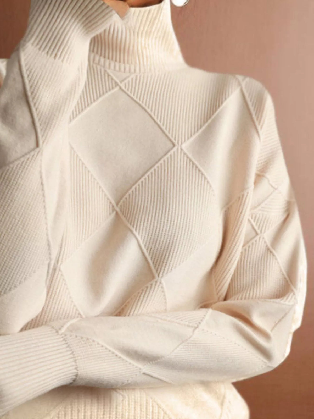 A close-up of a cream-colored knit turtleneck sweater with a textured diamond argyle pattern, shown on a model against a warm brown background.