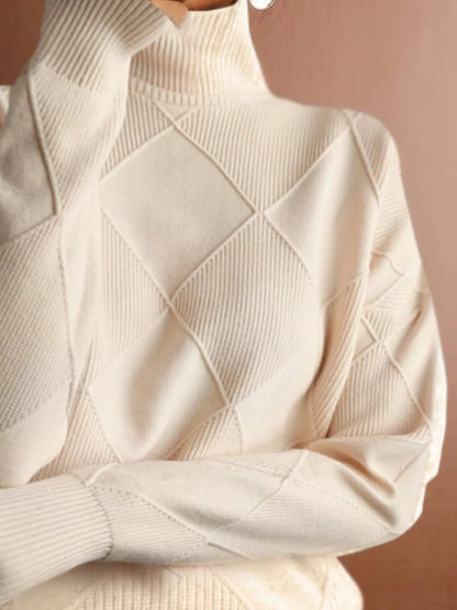A close-up of a cream-colored knit turtleneck sweater with a textured diamond argyle pattern, shown on a model against a warm brown background.