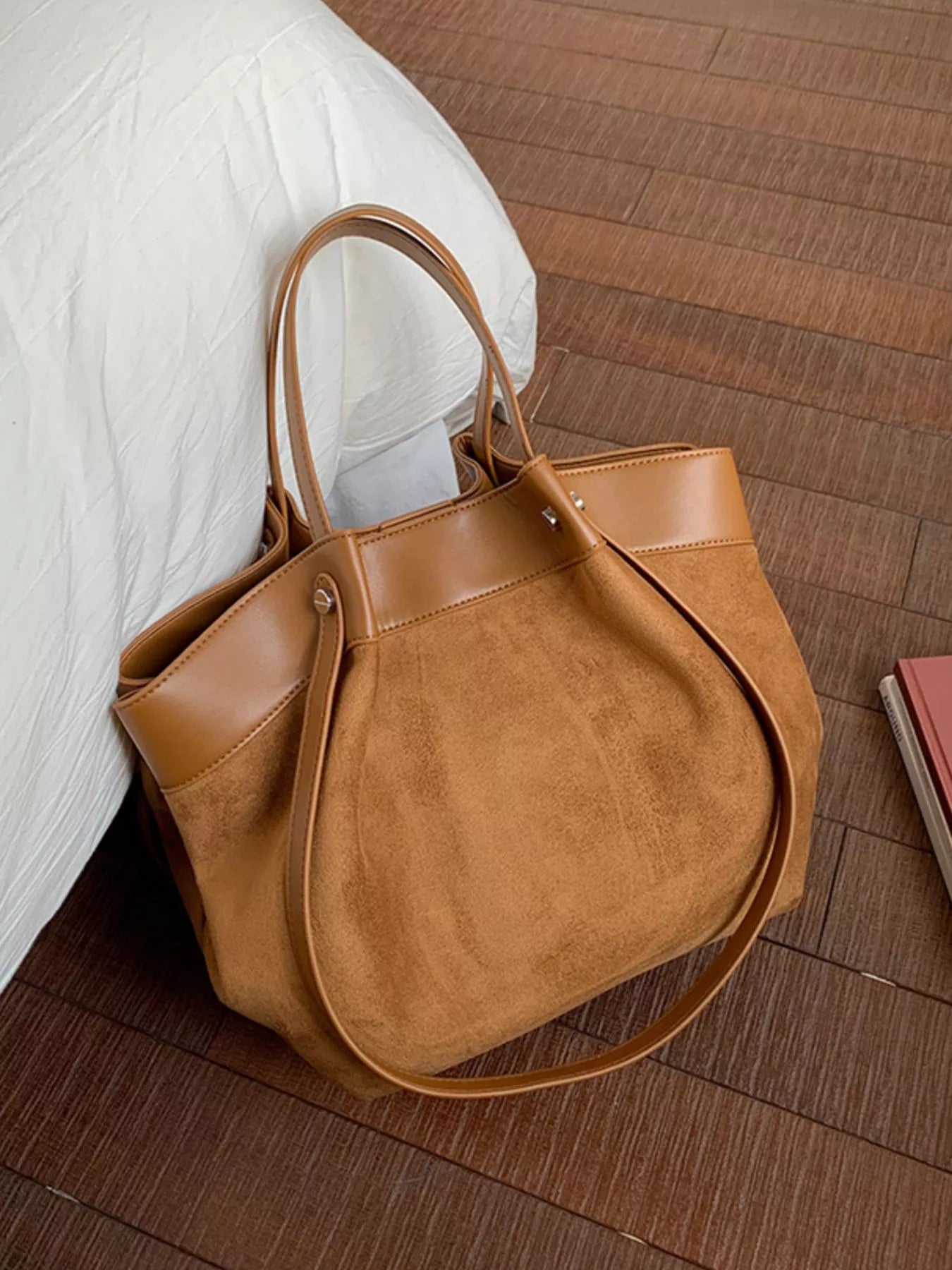 A brown suede and leather tote bag with double top handles, resting on a dark wood floor next to a white upholstered chair.