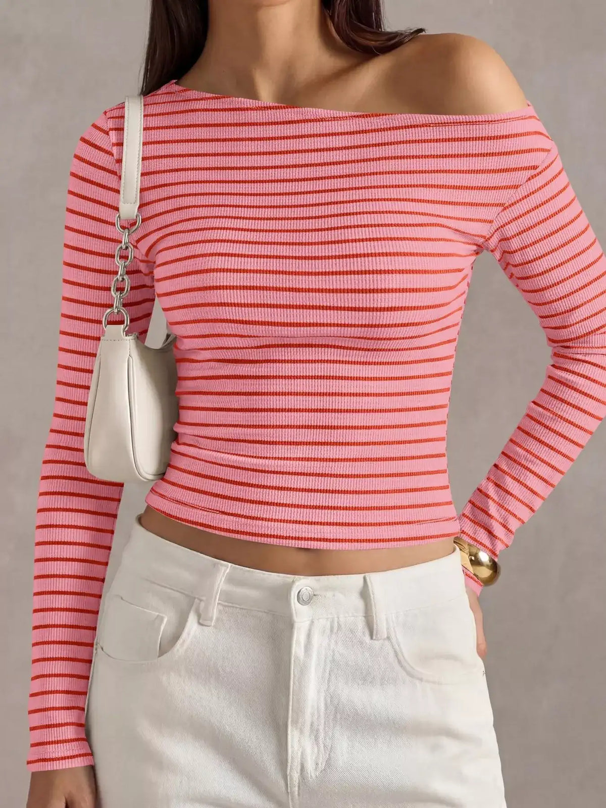 A woman wearing a pink and white horizontally striped off-the-shoulder long-sleeve crop top, paired with white jeans and a small white shoulder bag.