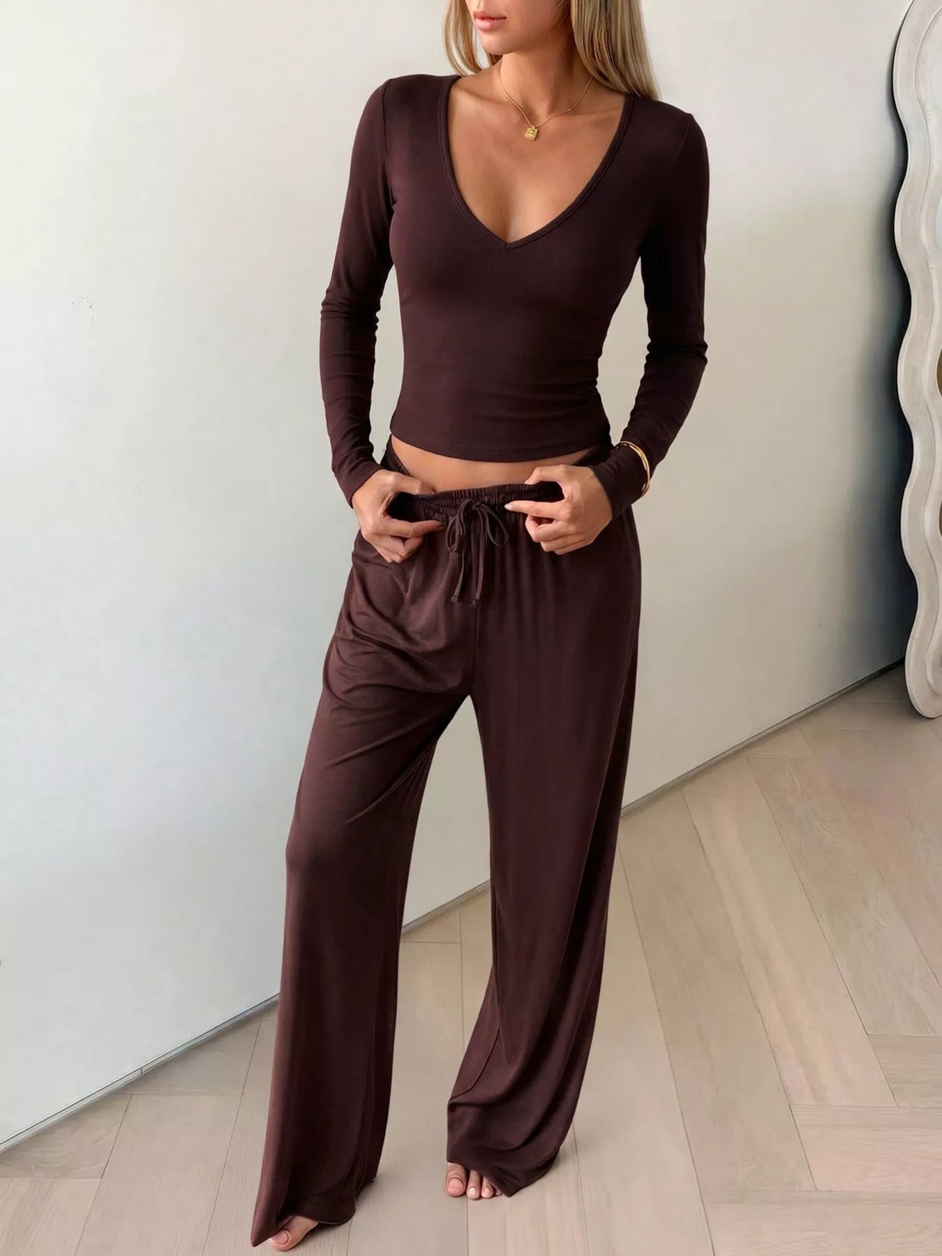 A woman modeling a dark brown two-piece loungewear set consisting of a long-sleeve V-neck crop top and matching wide-leg drawstring pants, standing on a light wood floor against a white wall.