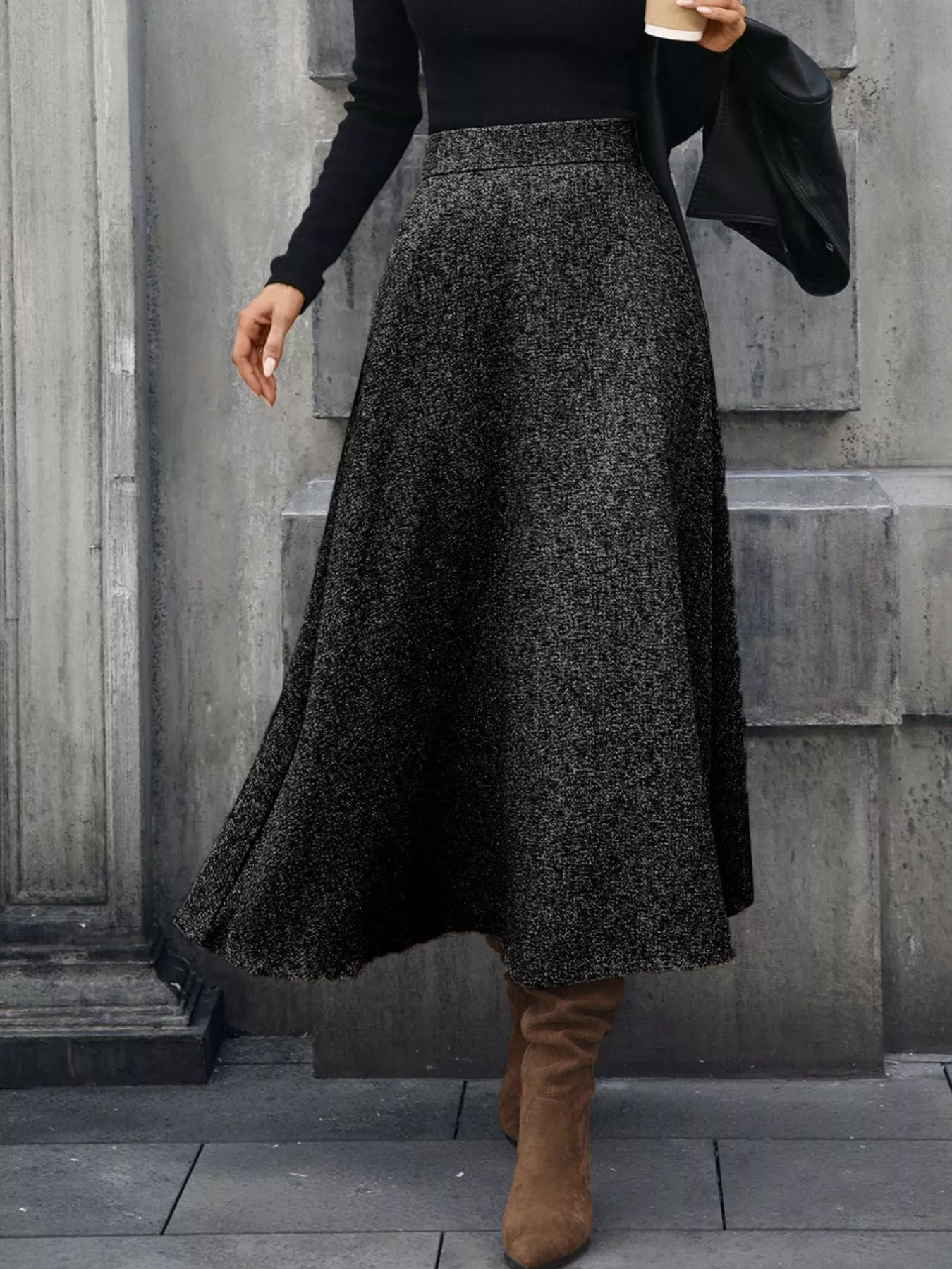 Person wearing a black and gray patterned skirt with a black top and brown boots against a gray wall.