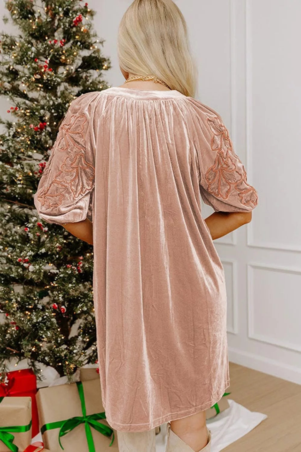 A woman viewed from behind wearing a dusty pink velvet tunic dress with lace puff sleeves, standing in front of a decorated Christmas tree and wrapped gifts.