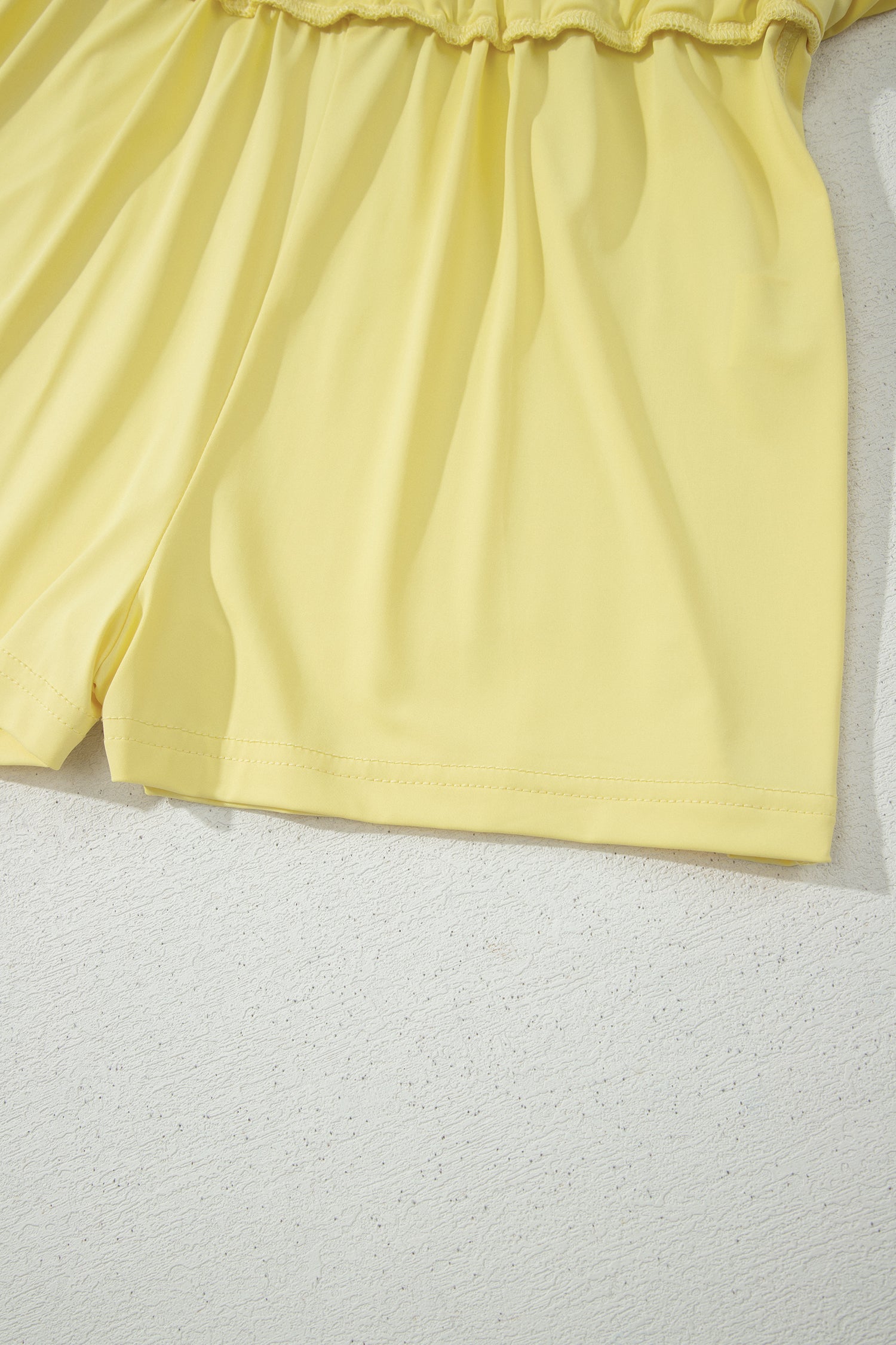 Close-up of a light yellow garment, showing the gathered waistband and upper leg portion, laid against a textured off-white background.