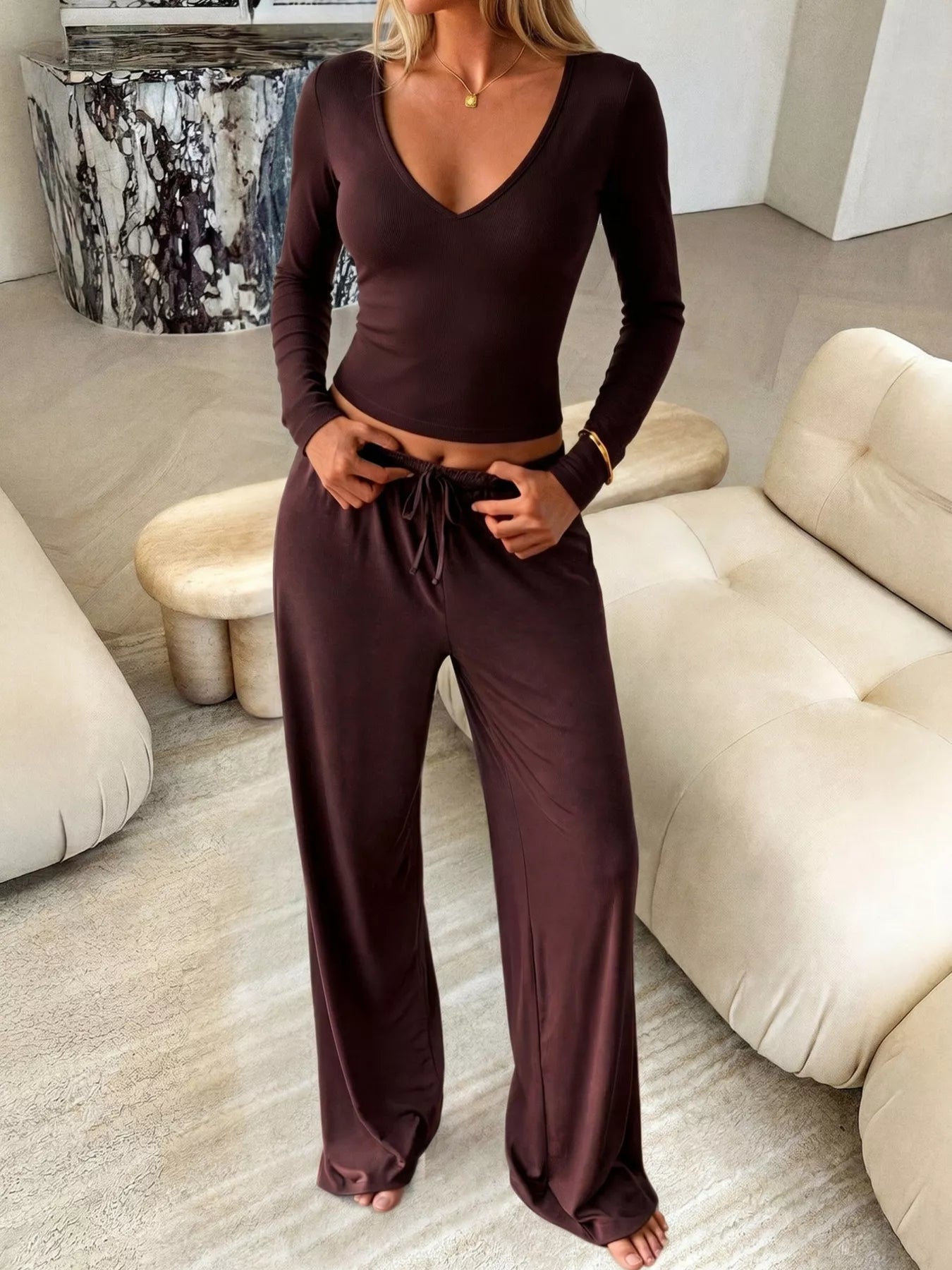A woman wearing a matching dark brown two-piece loungewear set consisting of a long-sleeve V-neck crop top and wide-leg drawstring pants, standing in a modern living room with beige sofas.