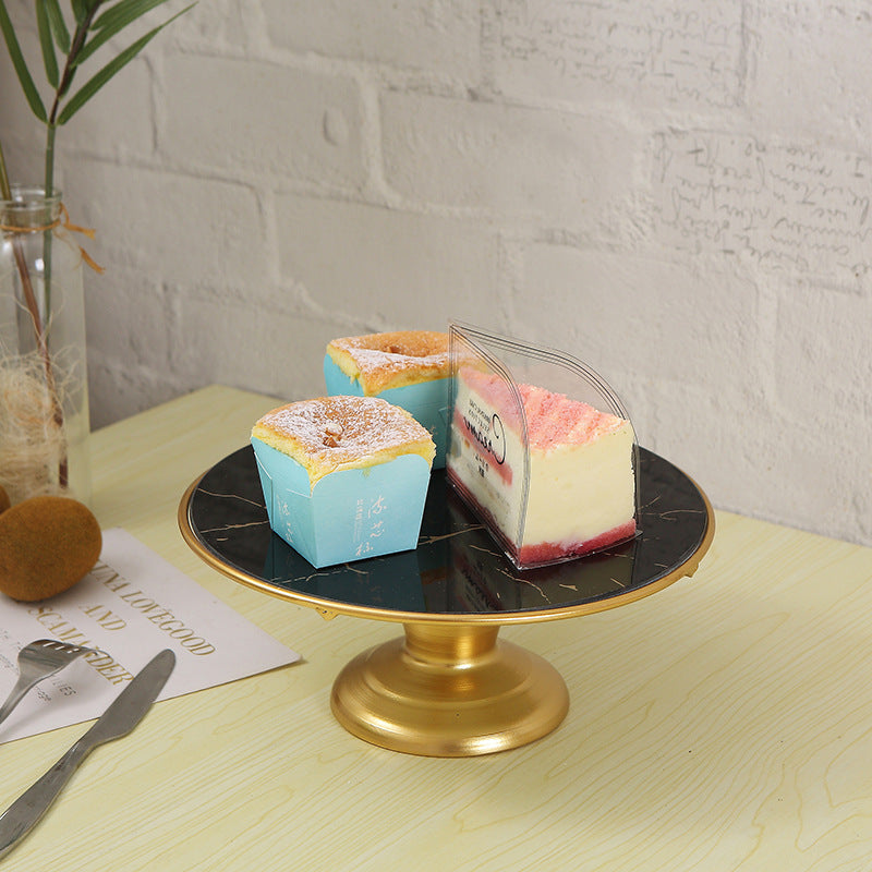 Three small cake slices in clear packaging on a black marble cake stand with a gold base, placed on a light wooden table.
