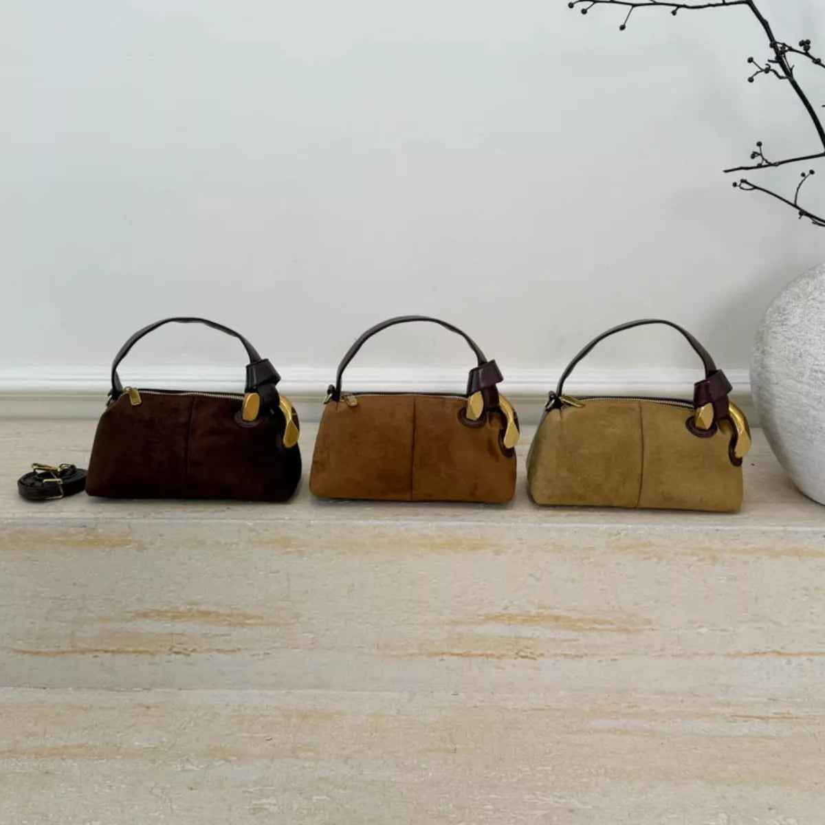 Three suede handbags in brown, tan, and beige on a light wooden floor.