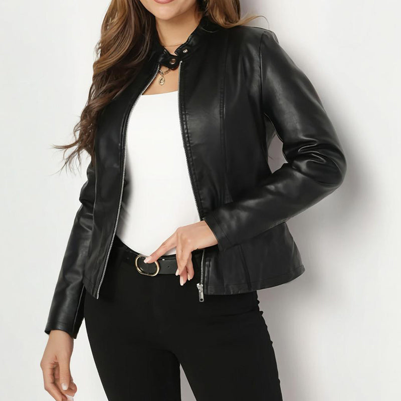 A woman wearing a black faux-leather moto jacket over a white top and black pants, standing against a plain light gray background.
