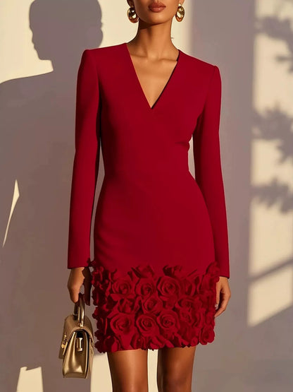 A woman models a fitted long-sleeve red mini dress with a deep V-neck and a rose-patterned hem, holding a small metallic gold handbag against a sunlit wall with leaf shadows.
