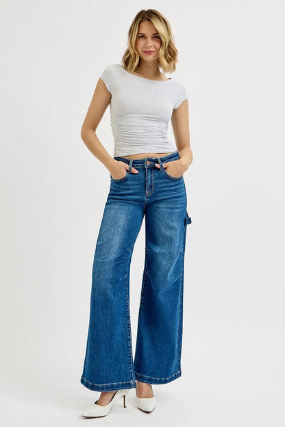 A woman modeling a light gray ribbed short-sleeve crop top and blue wide-leg denim jeans with white heels, standing against a plain light gray studio background.
