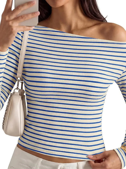 A woman wearing a blue and white horizontally striped off-the-shoulder long-sleeve ribbed top, holding a phone to take a selfie while wearing a small cream shoulder bag.
