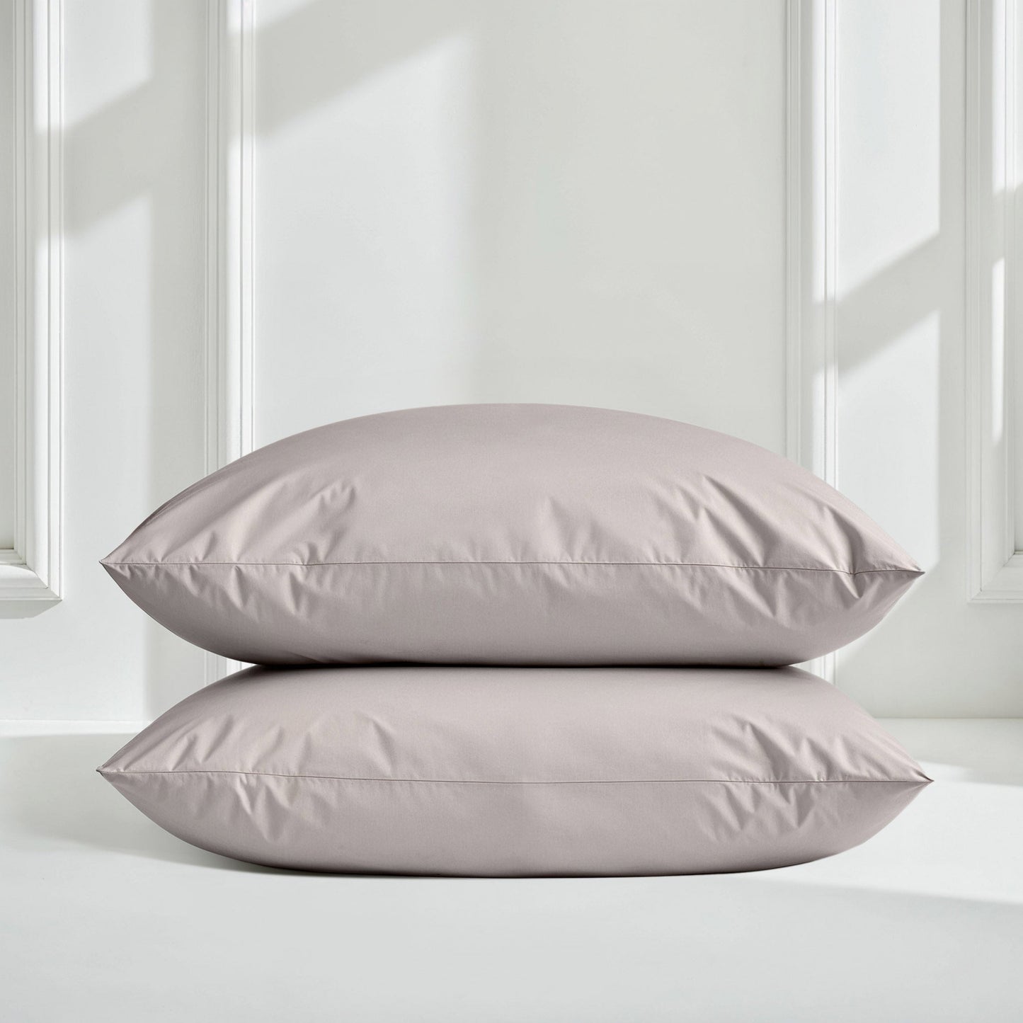 Two rectangular pillowcases stacked vertically on a white surface against a white paneled wall with soft window light shadows.