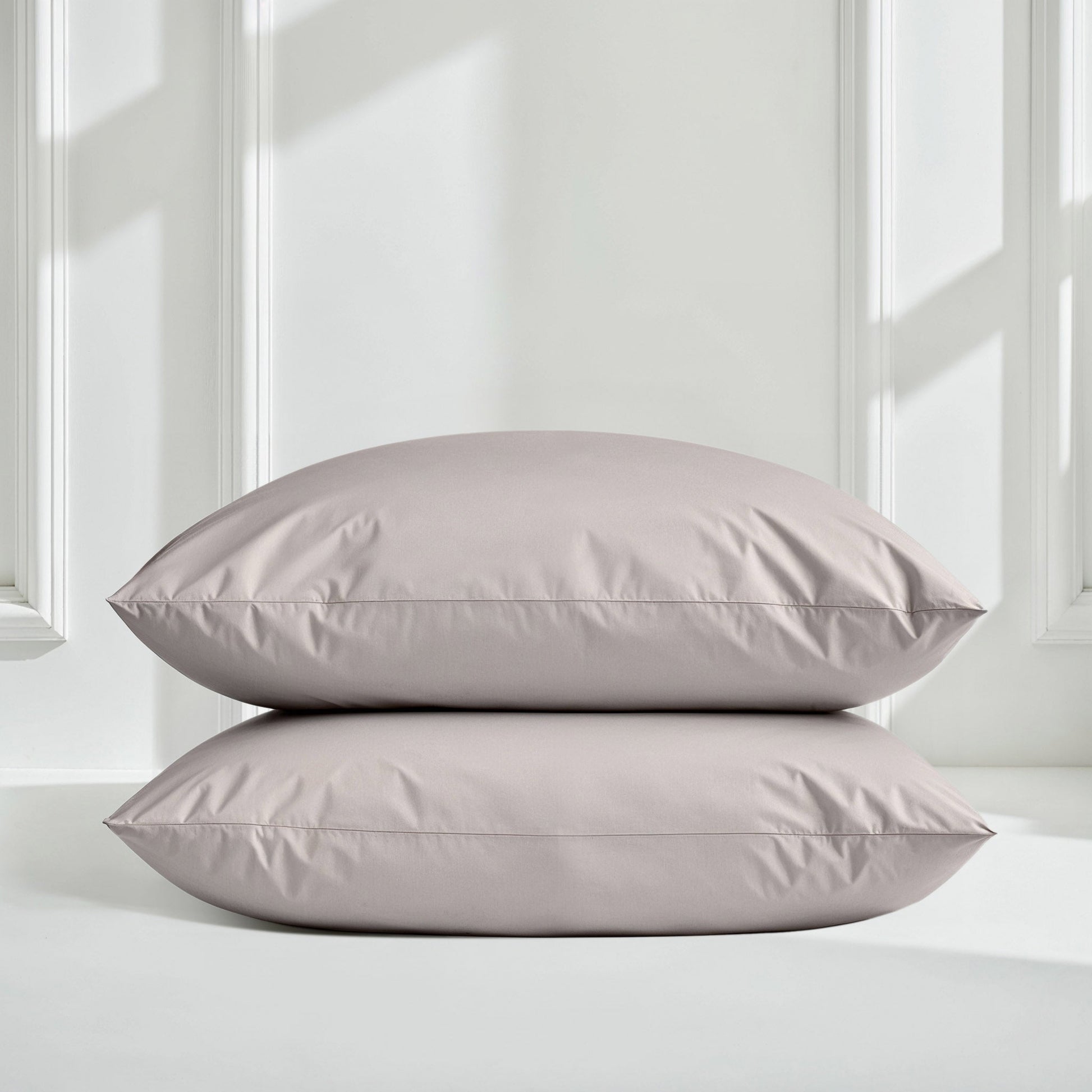 Two rectangular pillowcases stacked vertically on a white surface against a white paneled wall with soft window light shadows.