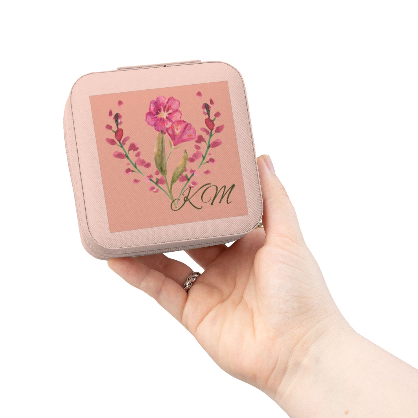 A hand holding a small square pink case with a floral illustration and the initials 'KM' on the lid, against a plain white background.