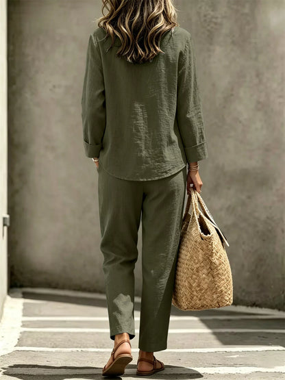 A woman shown from behind wearing an olive green linen two-piece set consisting of a long-sleeve button-up shirt and matching relaxed-fit trousers. She is carrying a large woven straw tote bag and wearing brown leather sandals, standing against a textured concrete wall.