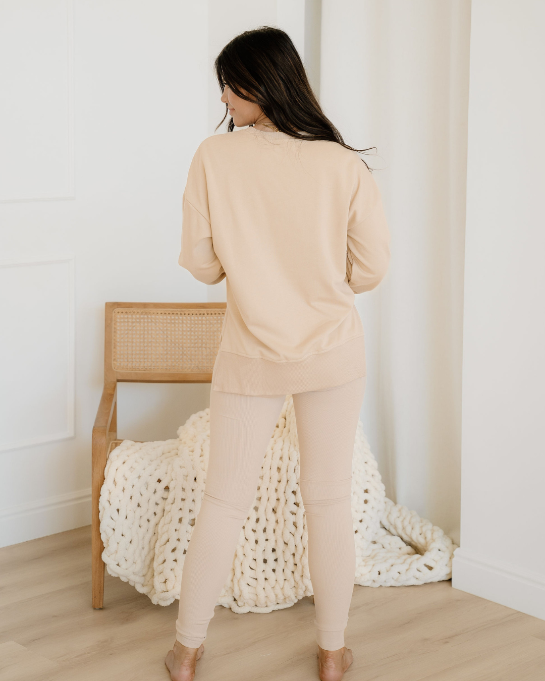 Woman in beige loungewear standing next to a wooden chair with a white blanket draped over it.